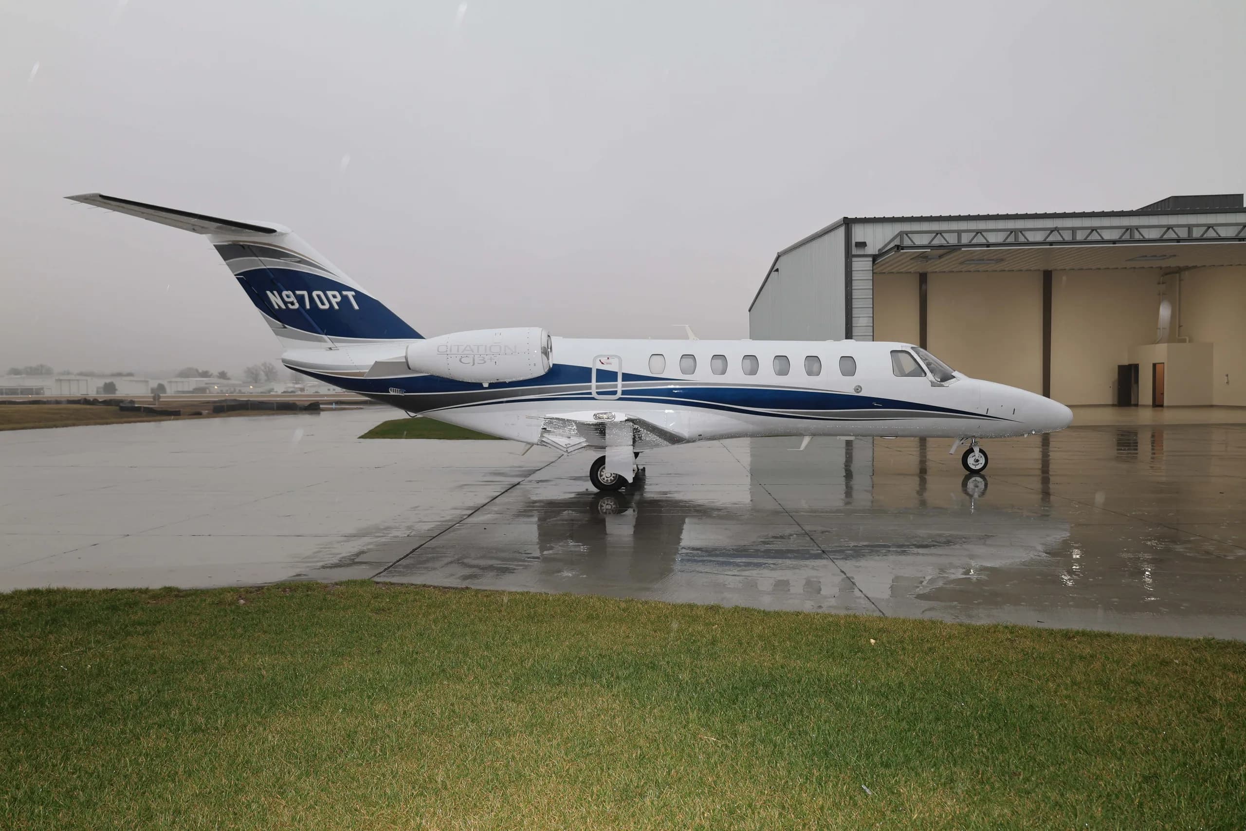 A white Cessna Citation M2 business jet with blue and grey stripes, registration N970PT, parked on a wet tarmac next to a hangar.