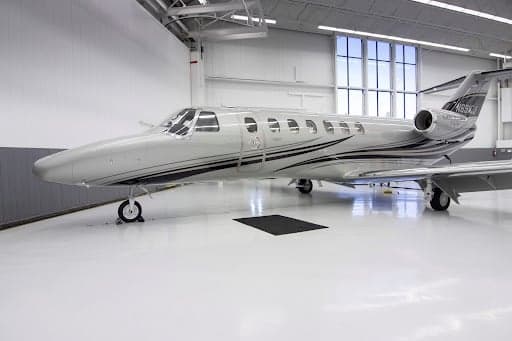 A silver Cessna Citation CJ3+ aircraft with black and grey stripes on the fuselage and tail, parked inside a brightly lit hangar.
