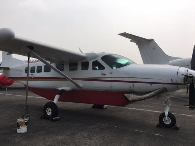 A white and red Cessna Caravan aircraft with a propeller, parked on an airfield, viewed from the front-side.