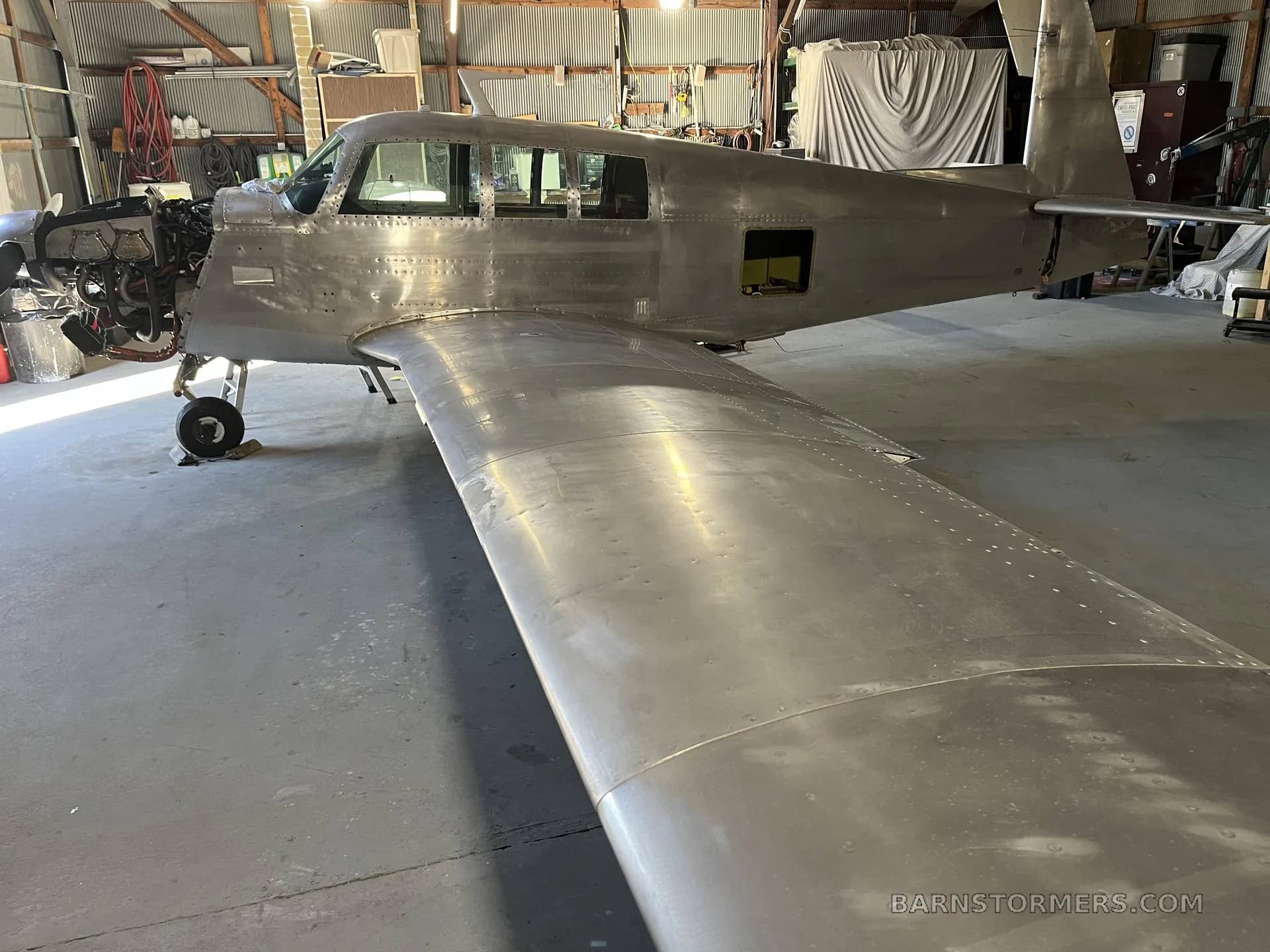 A bare metal aircraft, possibly a Mooney, undergoing restoration in a hangar, with its engine cowling removed.