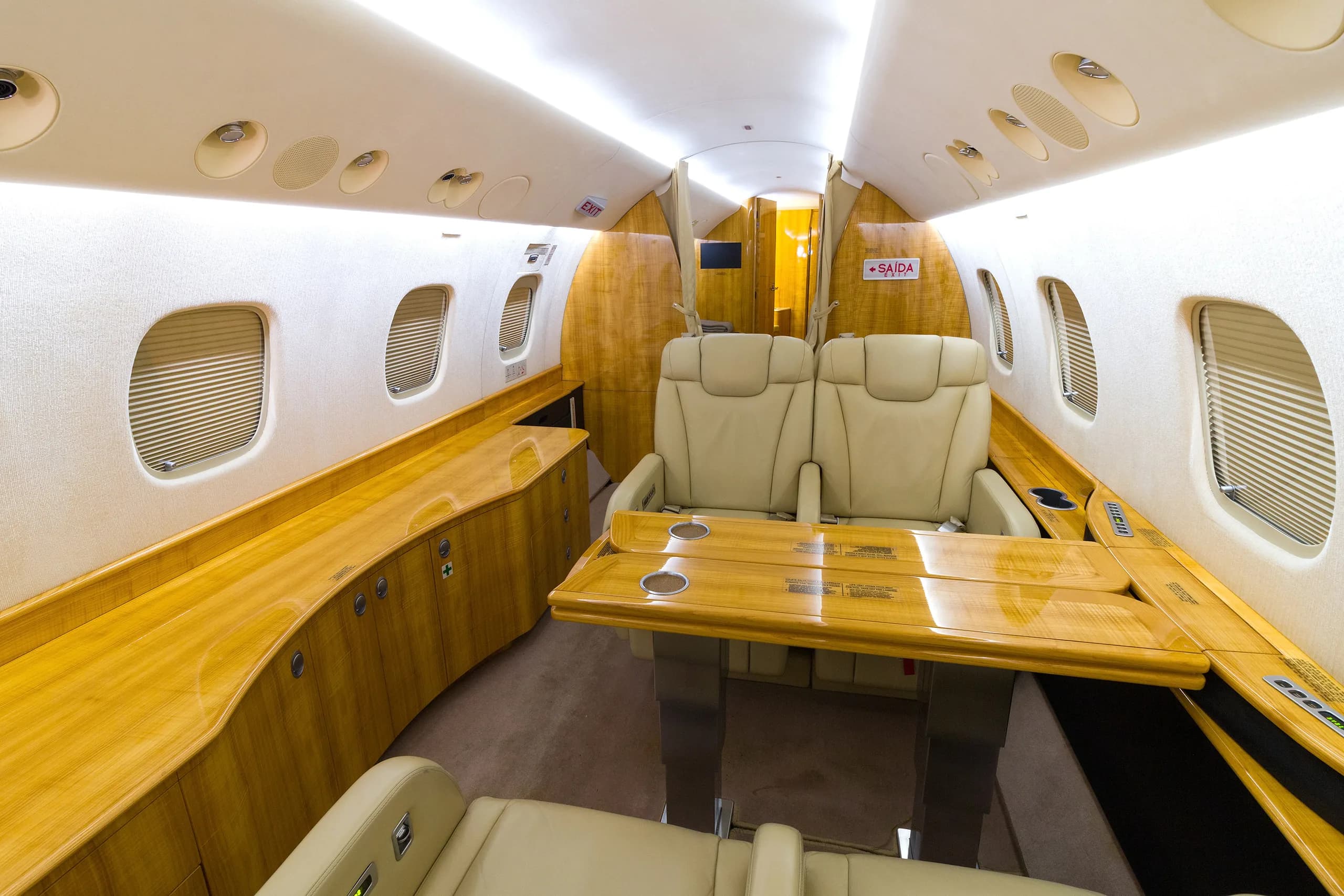 A private jet cabin interior with beige leather seats, a large polished wooden table, and extensive wooden cabinetry along the walls.