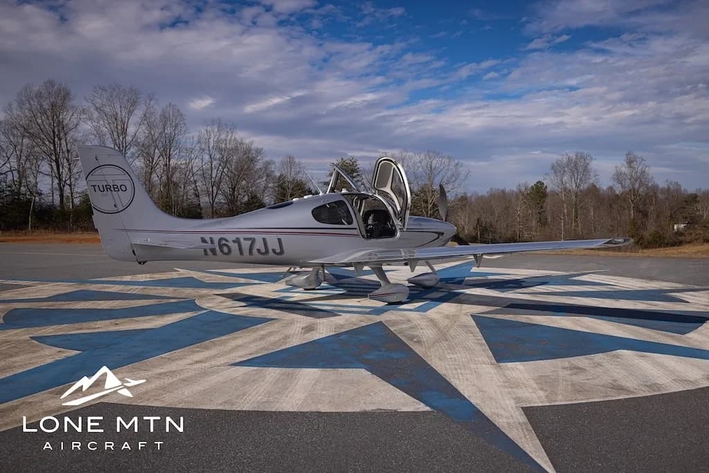 A silver Cirrus SR22T aircraft, N617JJ, with its doors open, parked on an airfield with trees in the background.