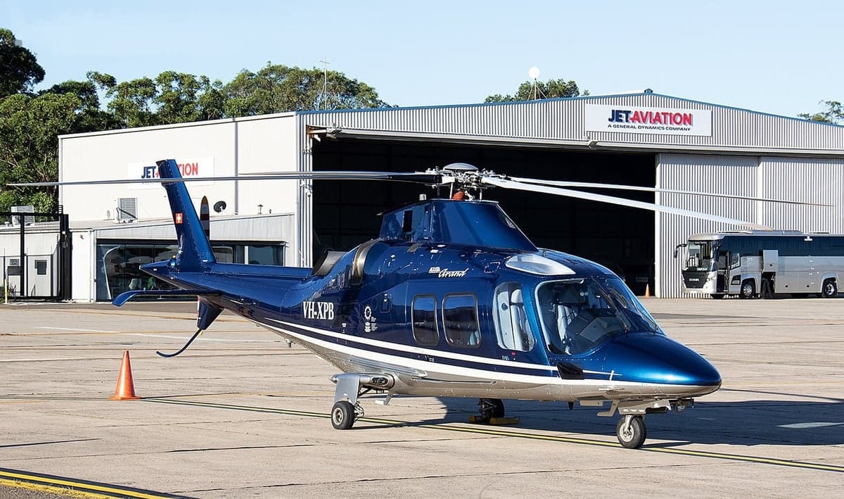 A blue Agusta Grand helicopter parked on the tarmac in front of a hangar.