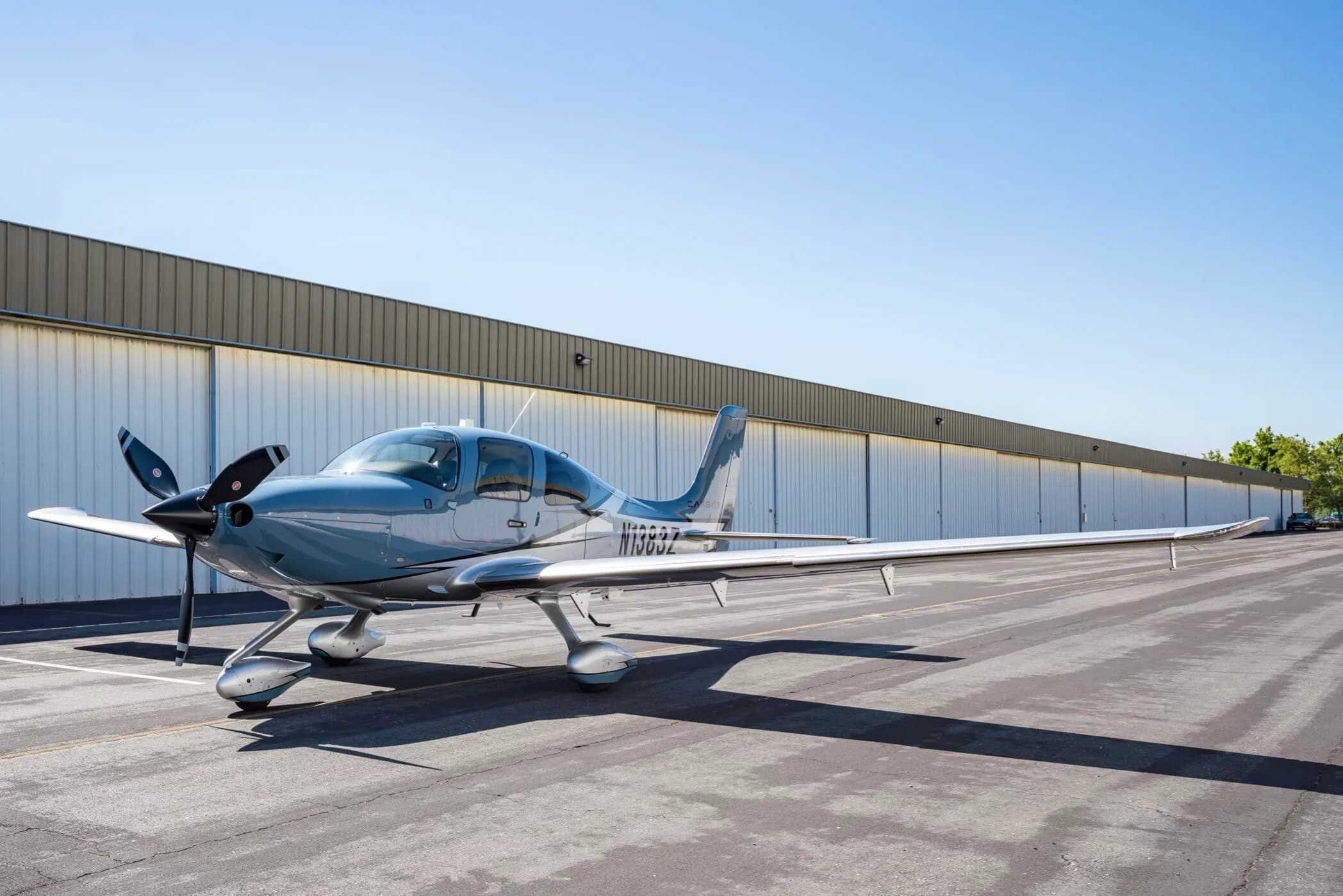 A light blue and silver Cirrus SR22T aircraft, N1383Z, parked on an tarmac in front of a long hangar.