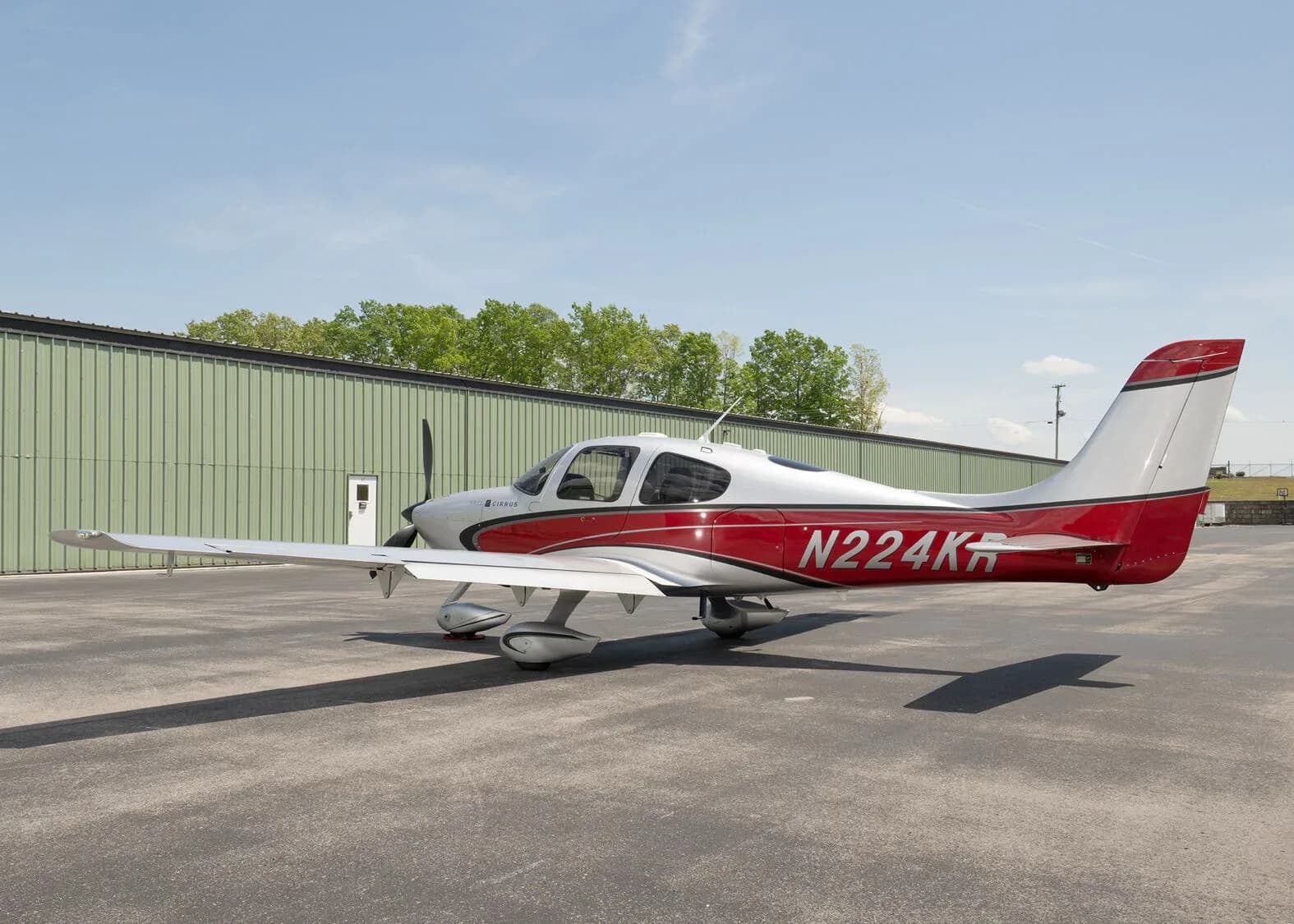 A silver and red Cirrus SR22 aircraft, N224KR, parked on an asphalt ramp next to a green hangar.