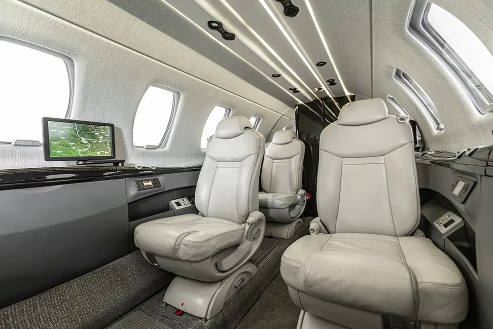 An angled view of the private jet's cabin, showcasing light grey seats, windows, and a screen displaying a map.