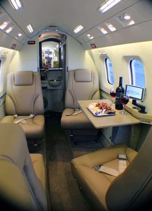 View of the aircraft cabin with tan leather seats, a table with wine and cheese, and a Petty logo on a screen.