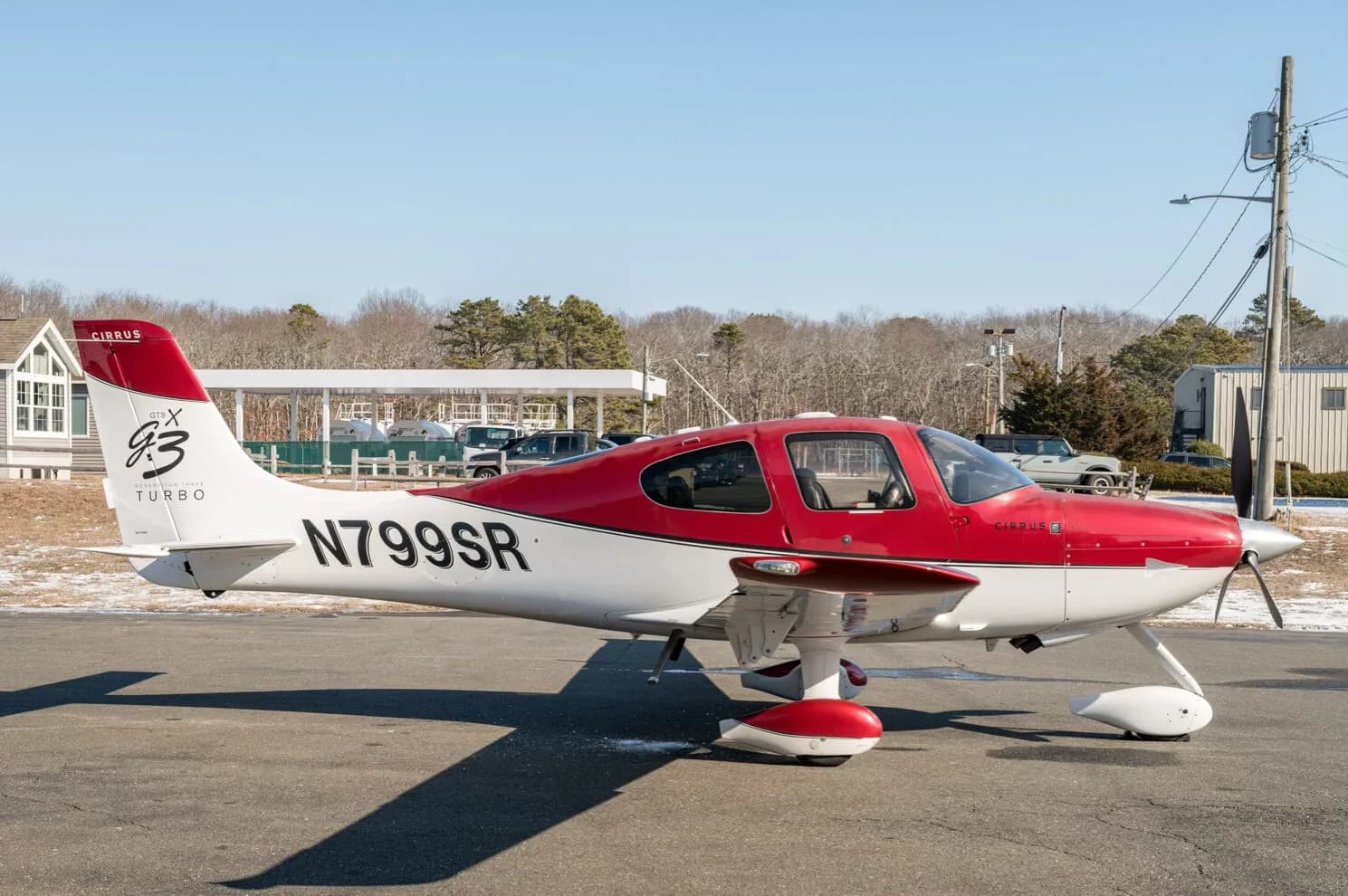 A red and white Cirrus SR22T aircraft, N799SR, parked on an tarmac with a building and trees in the background.