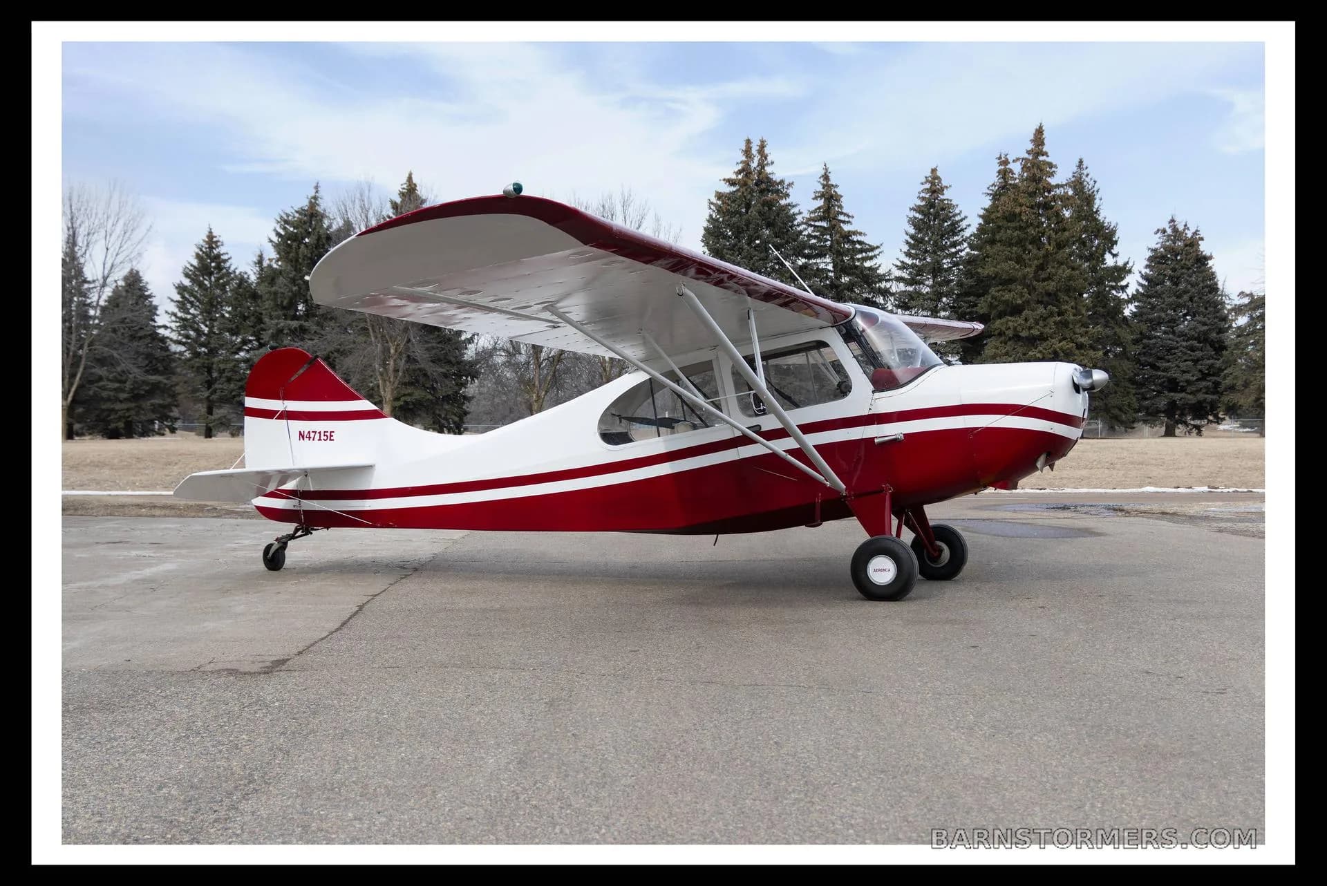 A red and white single-engine taildragger aircraft with registration N4715E, parked on an asphalt surface with pine trees in the background.