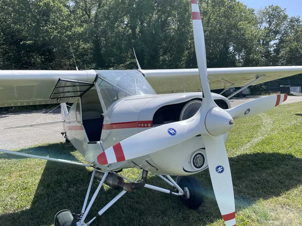 A front-quarter view of a white single-engine aircraft with red stripes, a three-bladed propeller, and an open door, parked on grass.