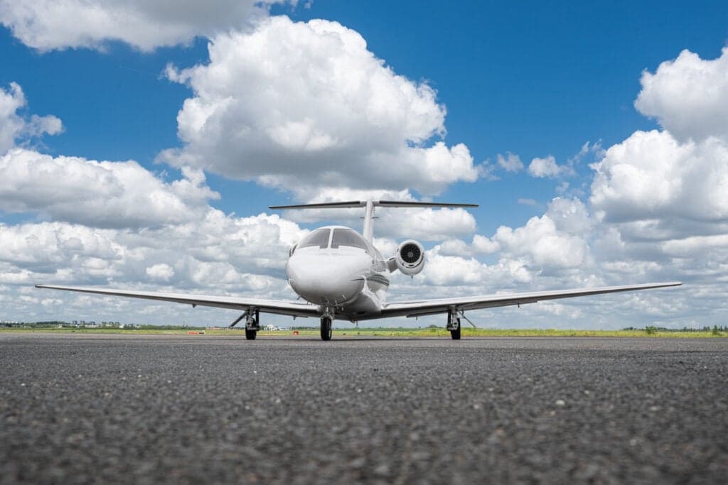 A white Cessna Citation CJ3 (D-IALI) private jet seen head-on from a low angle on an asphalt runway under a blue sky with white clouds.
