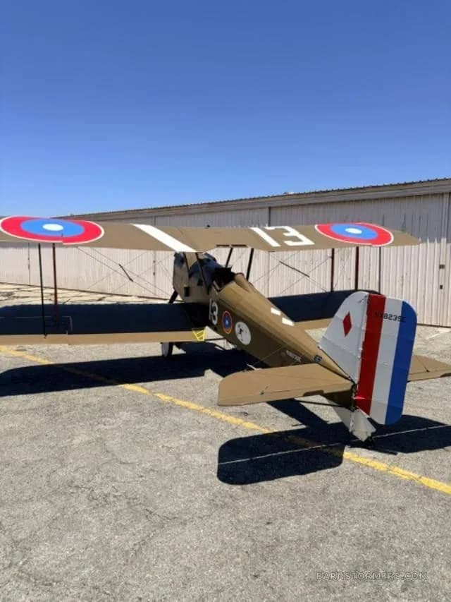 A rear-side view of a brown biplane with British roundels on the wings and a red, white, and blue rudder, parked on an asphalt apron.