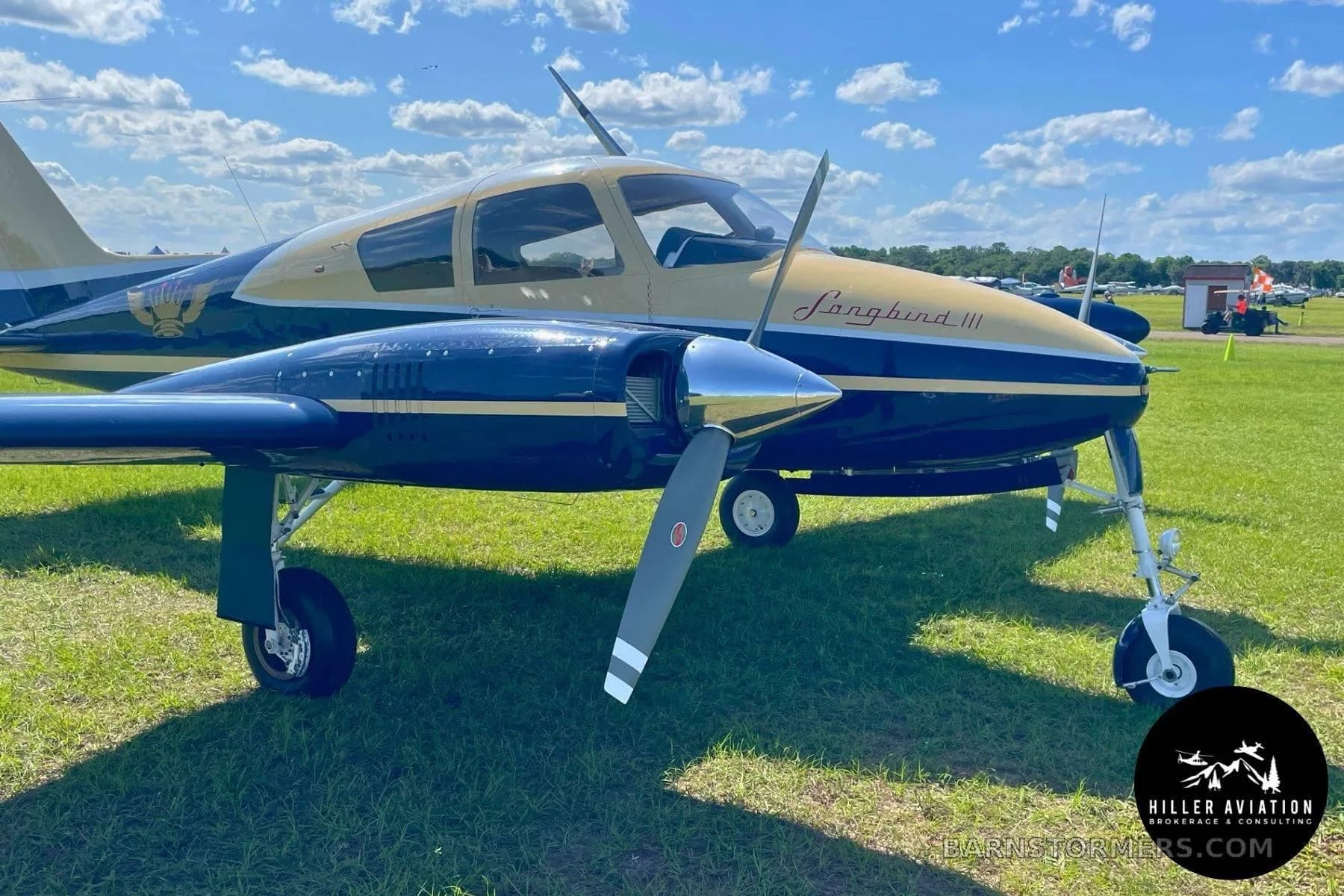 A close-up view of the front of a blue and gold twin-engine aircraft, "Longbird III", parked on a grassy field.