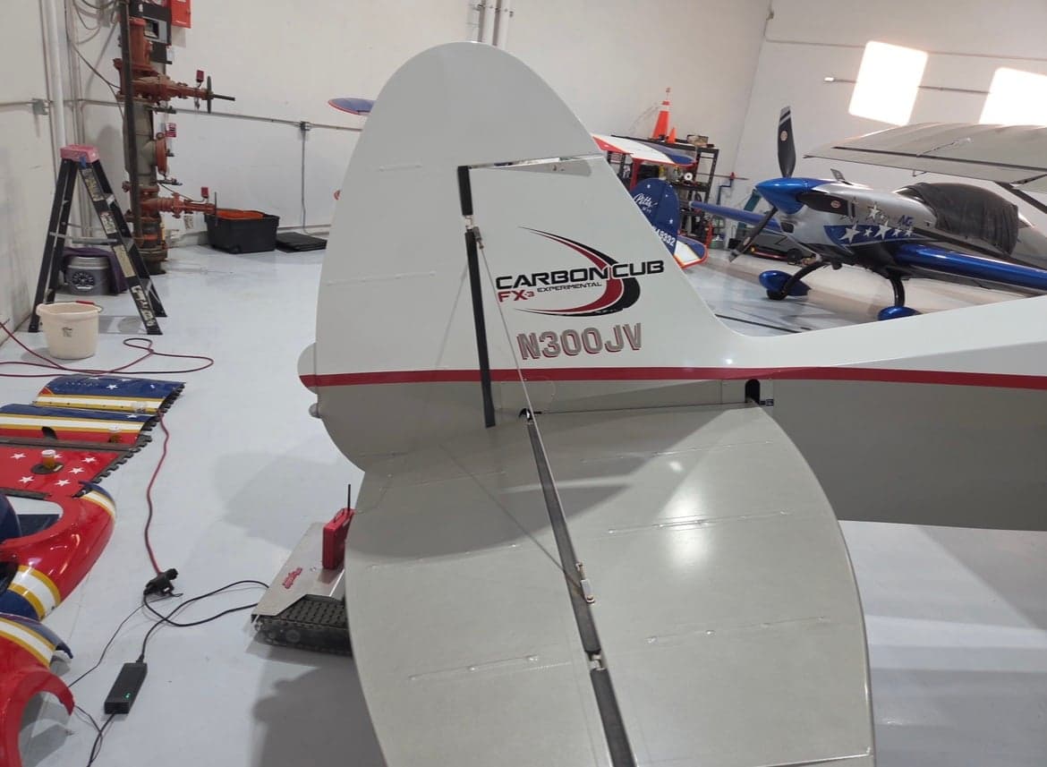 The tail of a white and red Carbon Cub aircraft, registration N300JV, is shown inside a hangar.