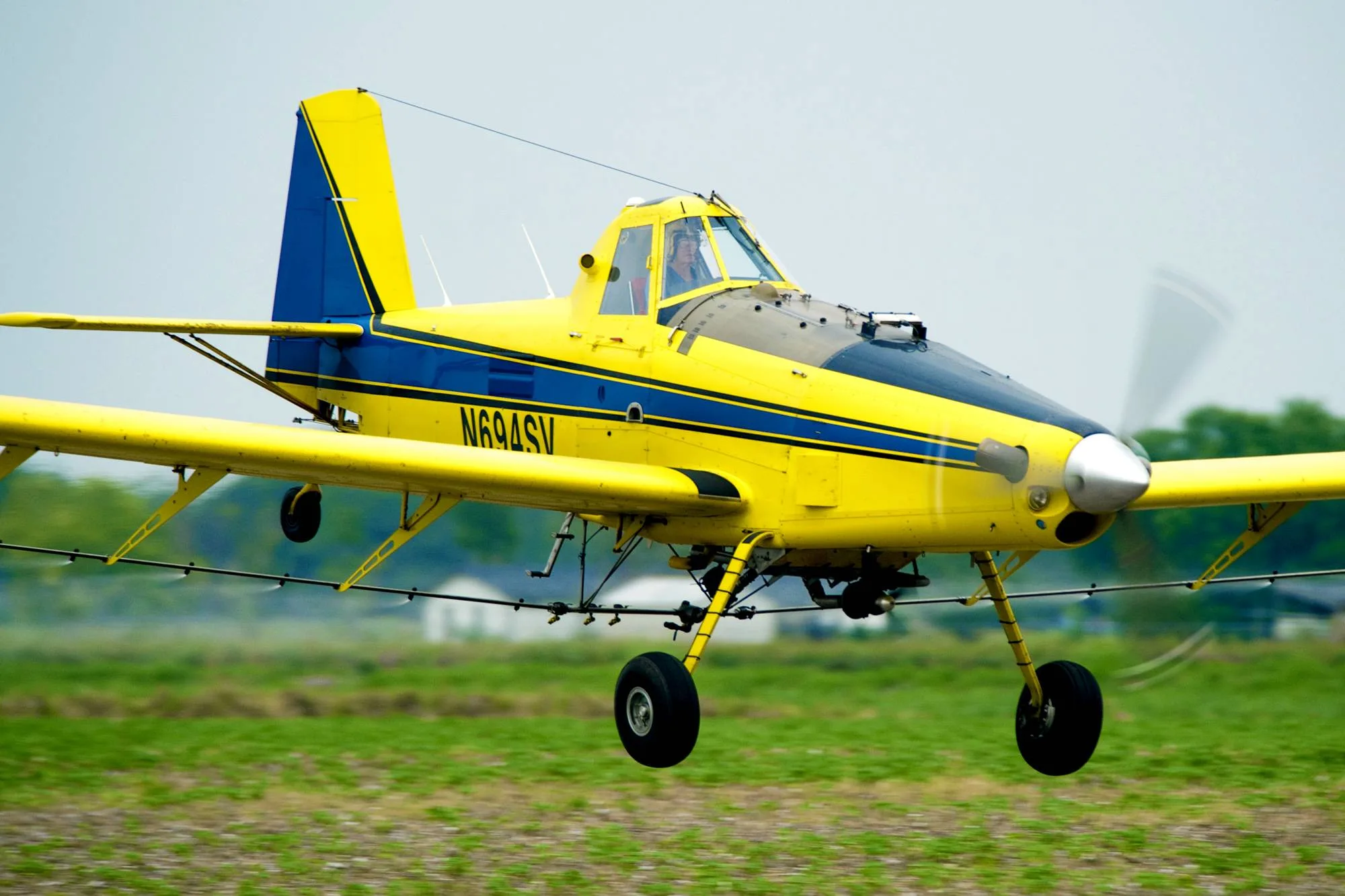 A yellow and blue agricultural aircraft, registration N694SV, flies low over a green field.