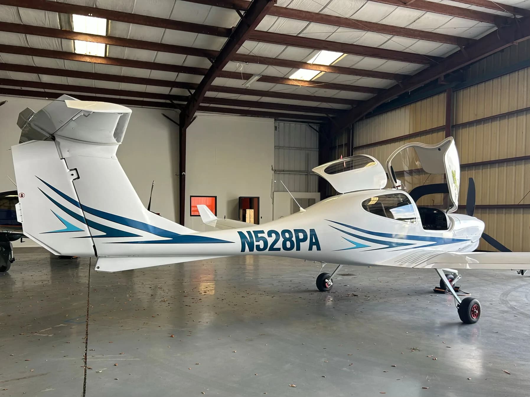 Rear-side view of a white Diamond DA40 aircraft with blue and light blue stripes, registration N528PA, parked in a hangar with its canopy and rear hatch open.