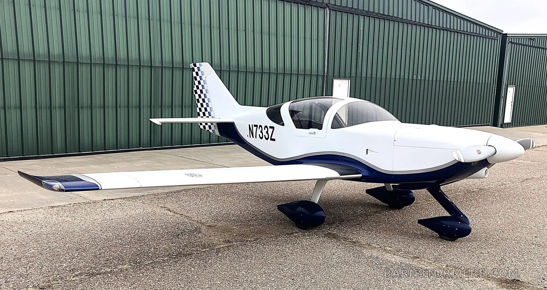 A white and blue single-engine aircraft with registration N733Z parked on an airfield in front of a green hangar.