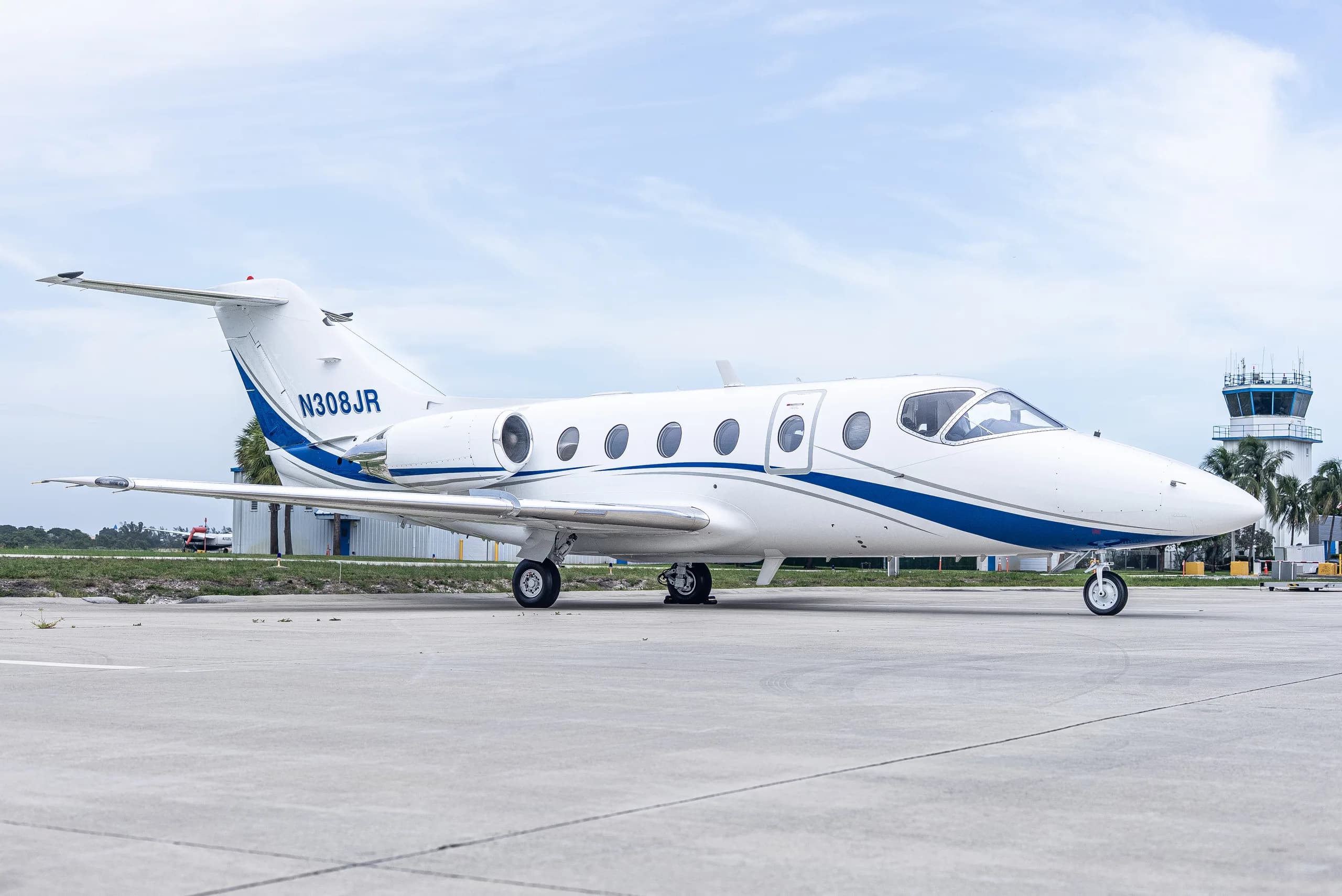 A white business jet with blue stripes, registration N308JR, is parked on an airport tarmac with a control tower in the background.