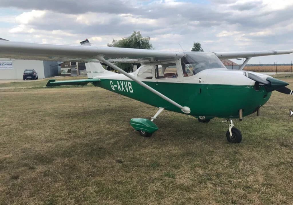 A white and green Cessna 172 (G-AXVB) parked on a grassy airfield under a cloudy sky.