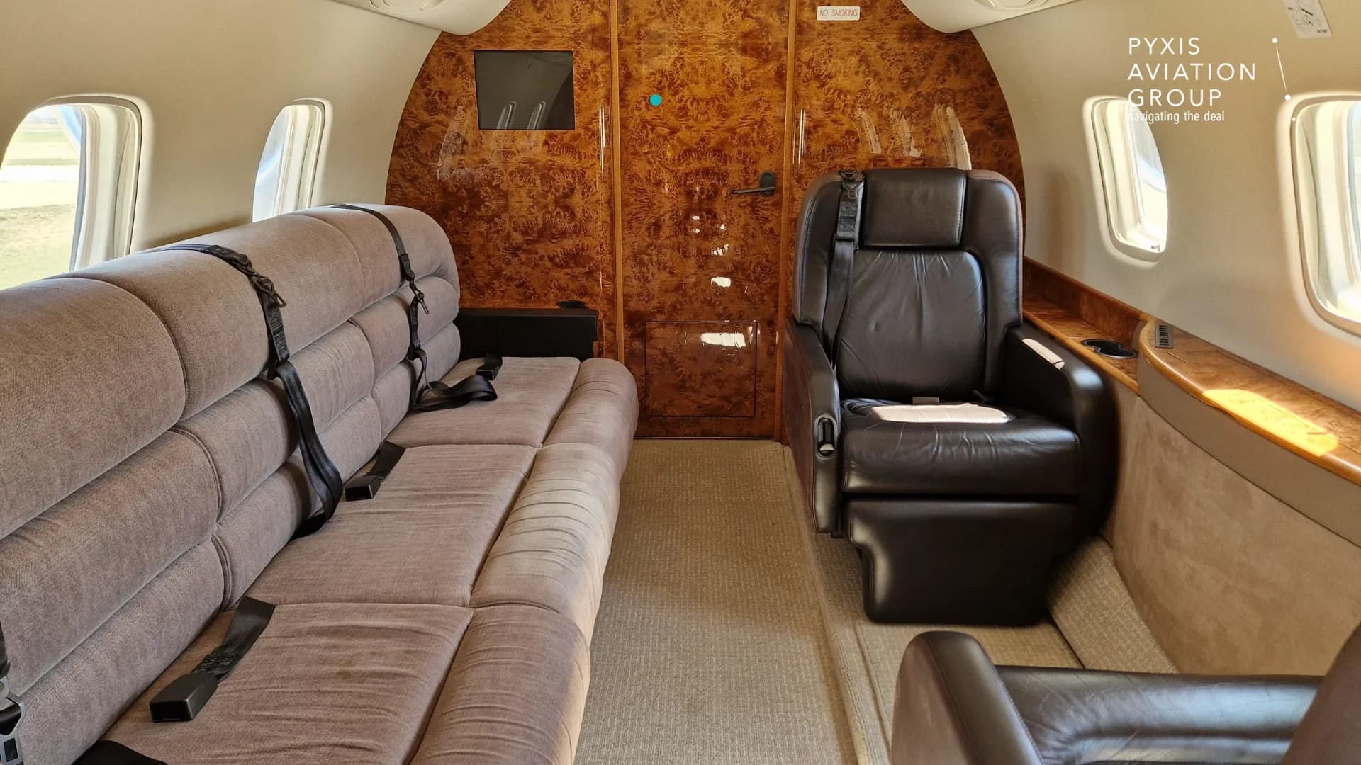 The aircraft cabin interior showing a long beige sofa-style seat on the left and a single dark leather seat on the right, with wood paneling.
