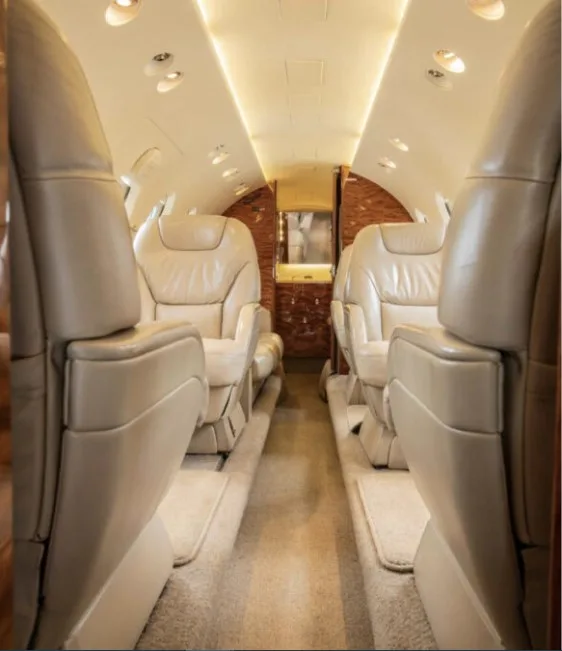 A view down the aisle of a business jet's cabin, featuring rows of plush beige leather seats and wood paneling.