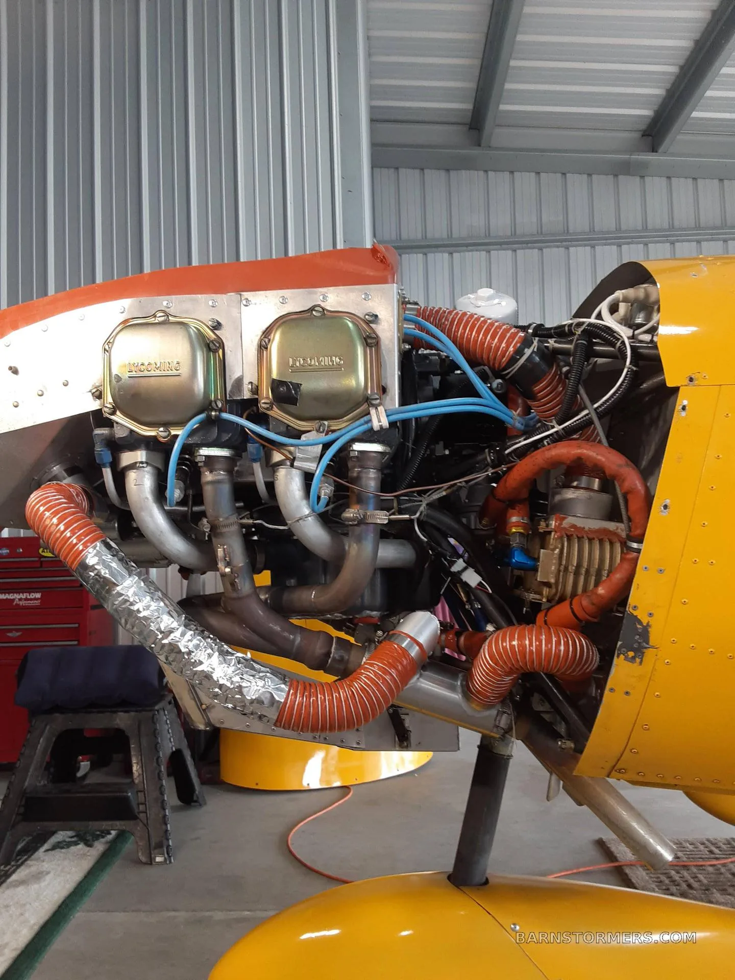 The exposed engine compartment of a yellow aircraft, showing the engine, exhaust pipes, and various hoses in a hangar setting.