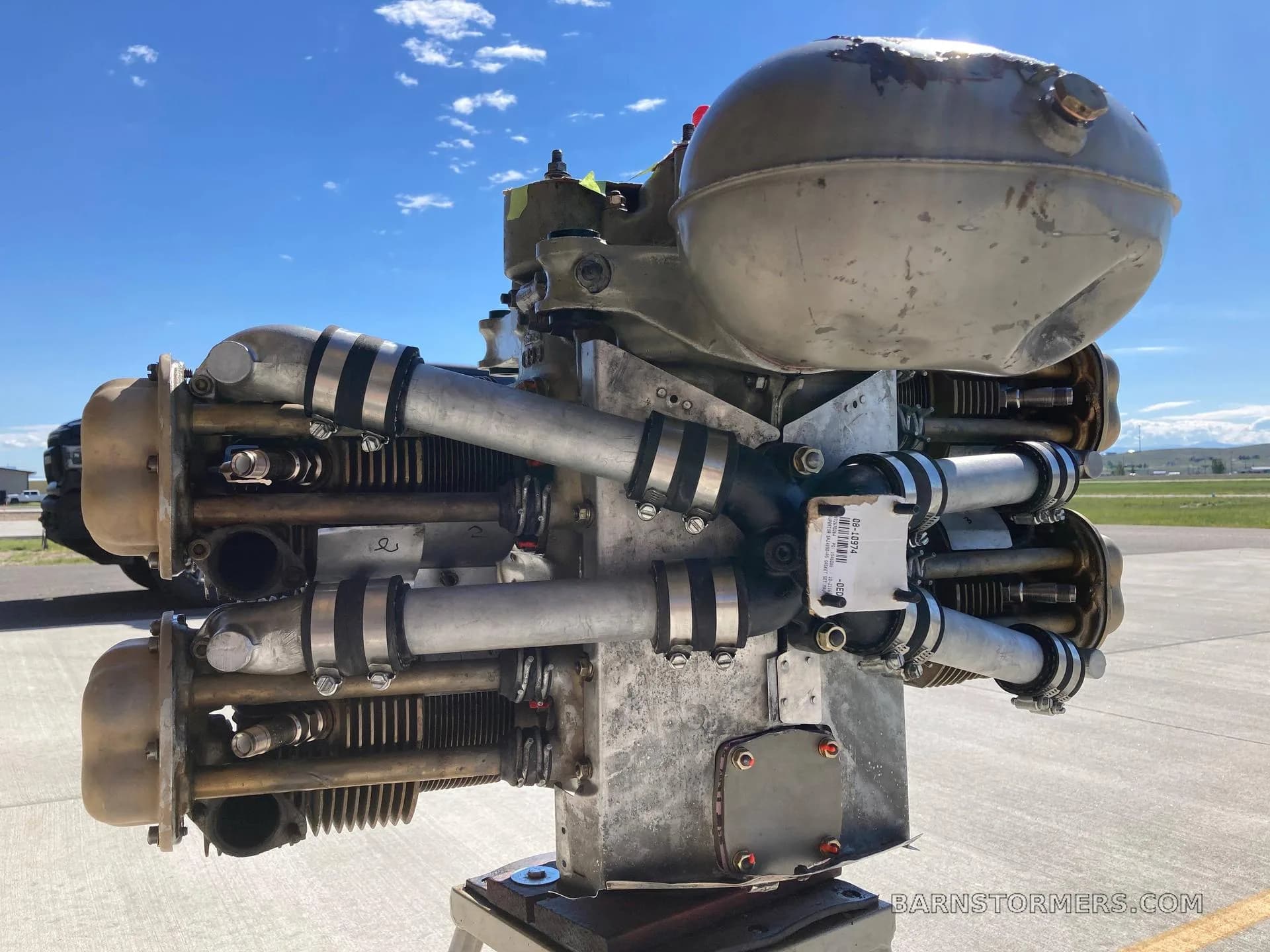 A grey aircraft engine with intake pipes on an airfield tarmac under a blue sky.