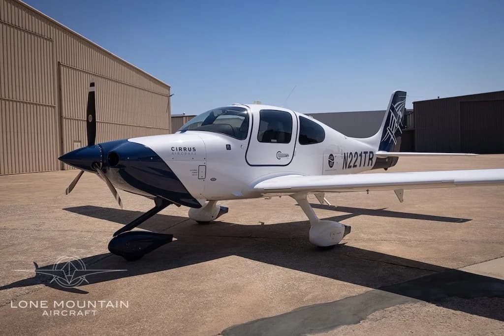 A three-quarter front view of the white Cirrus SR22 aircraft with a dark blue nose and tail, registration N221TR, parked on an airport tarmac.