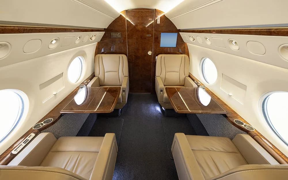 The spacious cabin interior of a private jet, featuring two sets of facing beige leather seats with polished wood tables.