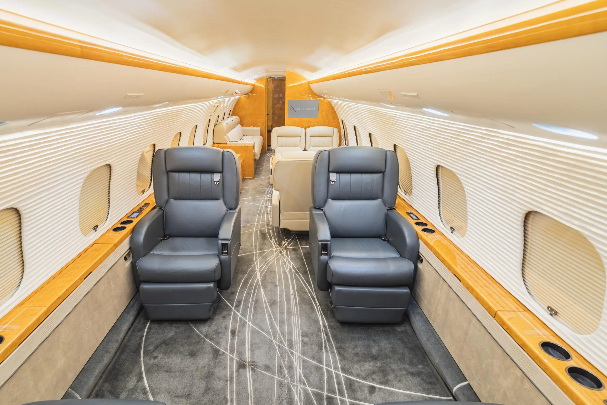 The luxurious cabin interior of a business jet, featuring two grey leather seats in the foreground and a light wood and cream color scheme.