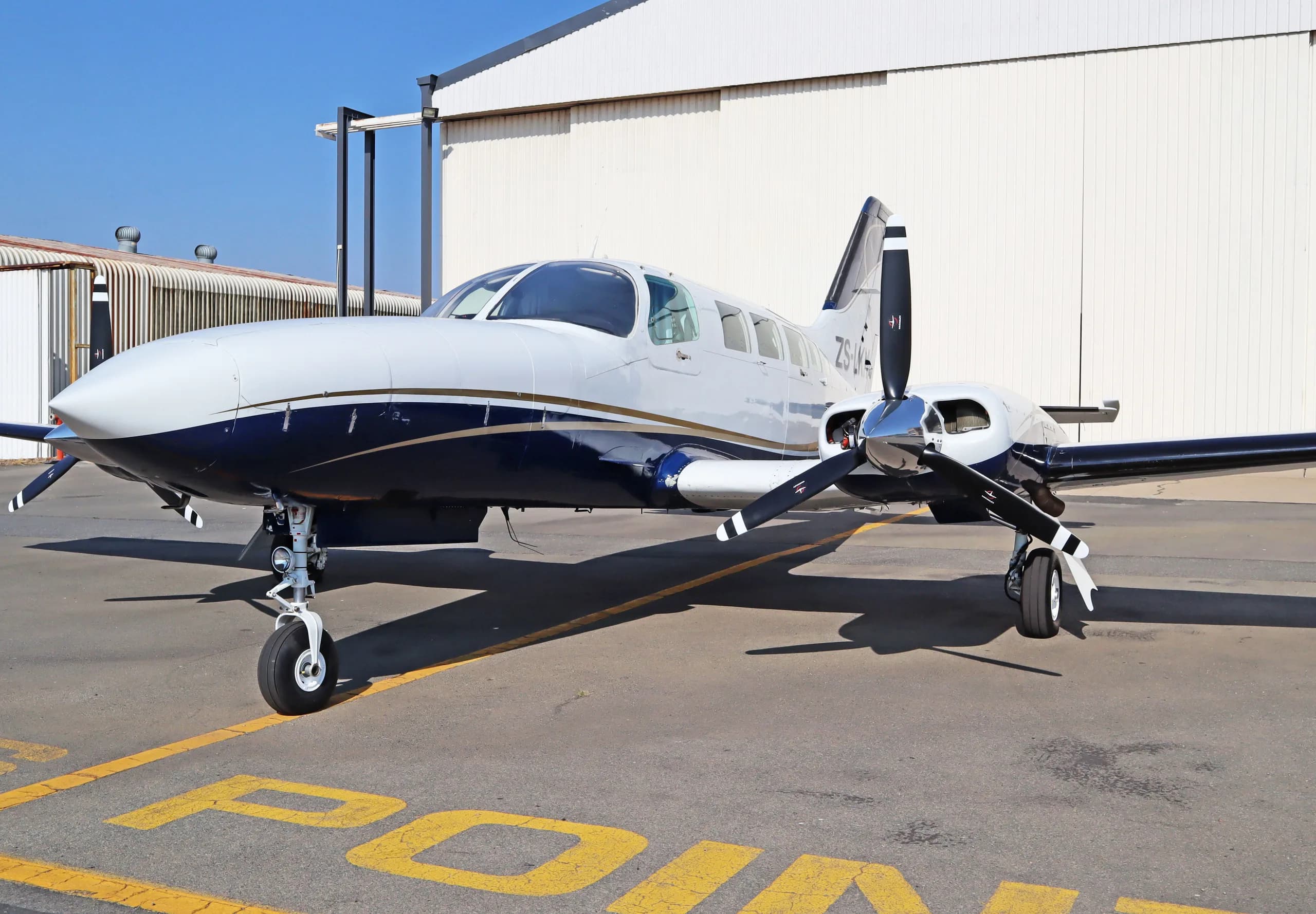 A white and dark blue twin-engine propeller aircraft with a gold stripe parked on an asphalt tarmac next to a hangar.