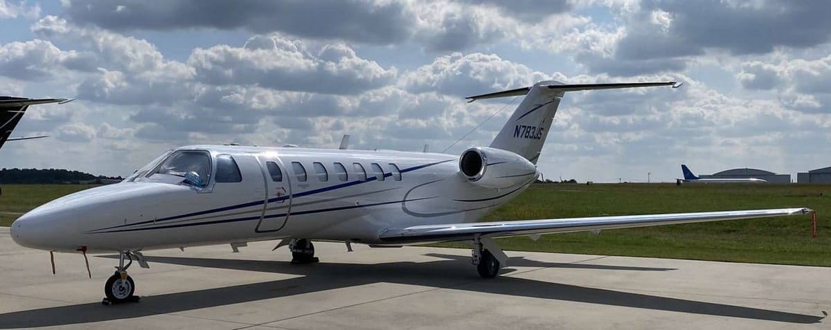 A Cessna Citation CJ3 with a white and blue paint scheme is parked on the tarmac under a cloudy sky.