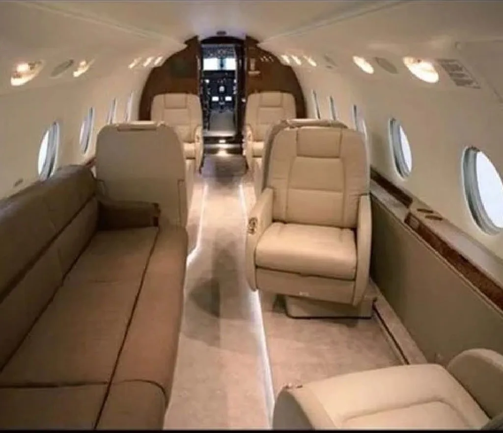 The luxurious interior cabin of a private jet, featuring beige leather seats and a sofa, with a view towards the cockpit.