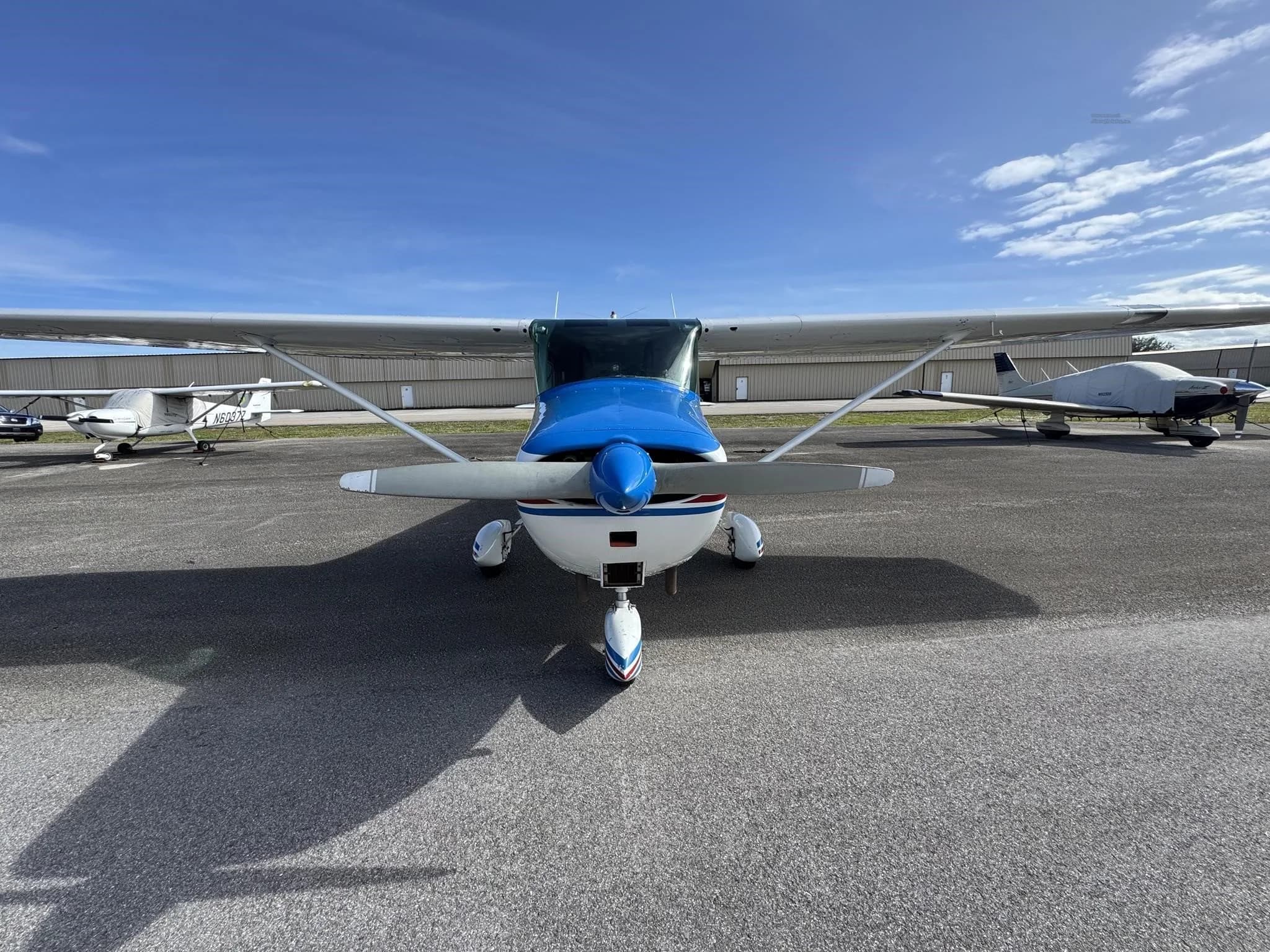 Front view of a white and blue Cessna 172 aircraft with a blue engine cover, parked on an airfield.