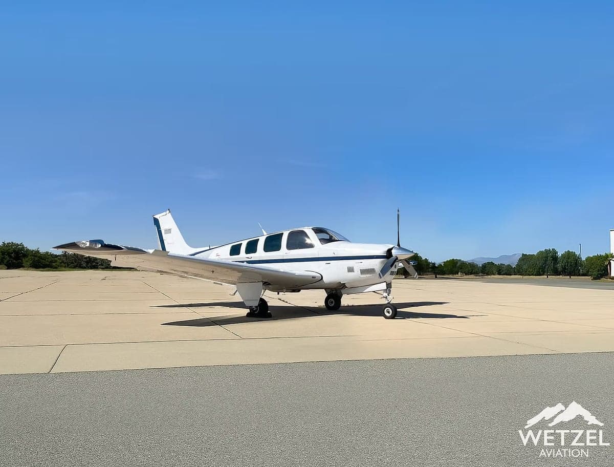 A white and blue Beechcraft Bonanza sits on the tarmac under a clear blue sky.