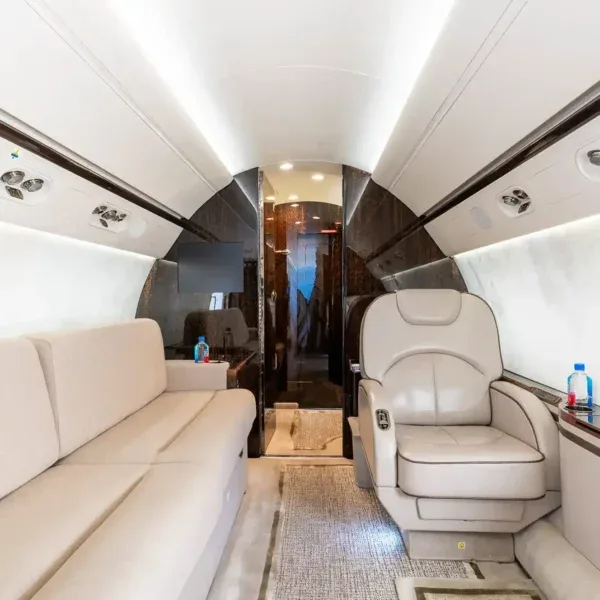 A section of the aircraft cabin interior featuring a long, light-colored sofa and a matching single seat, with dark wood accents.