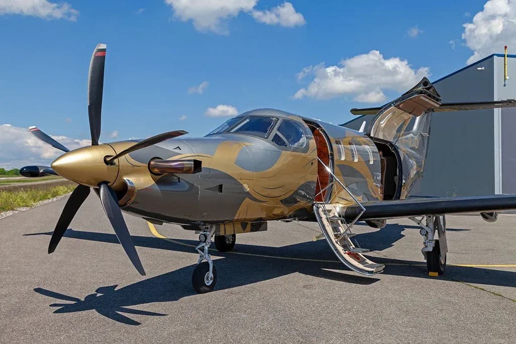 A front-side view of a silver and gold Pilatus PC-12 aircraft with its doors open, parked on an asphalt runway next to a hangar.