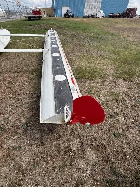 An end view of a white aircraft float with a red tail section, on the grass near a hangar.