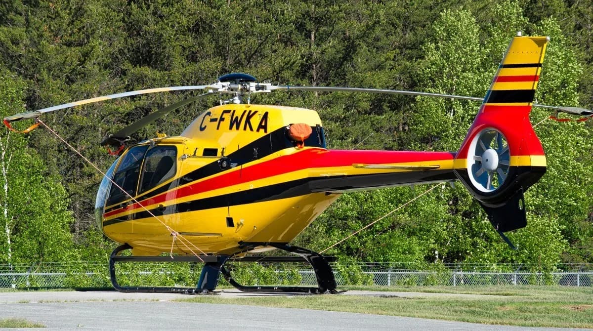 A yellow helicopter with red and black stripes, registration C-FWKA, is parked on a paved surface with trees in the background.