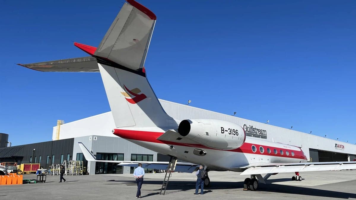 A rear-quarter view of a red and white Gulfstream G650 business jet parked in front of a hangar, showing its tail and engine.