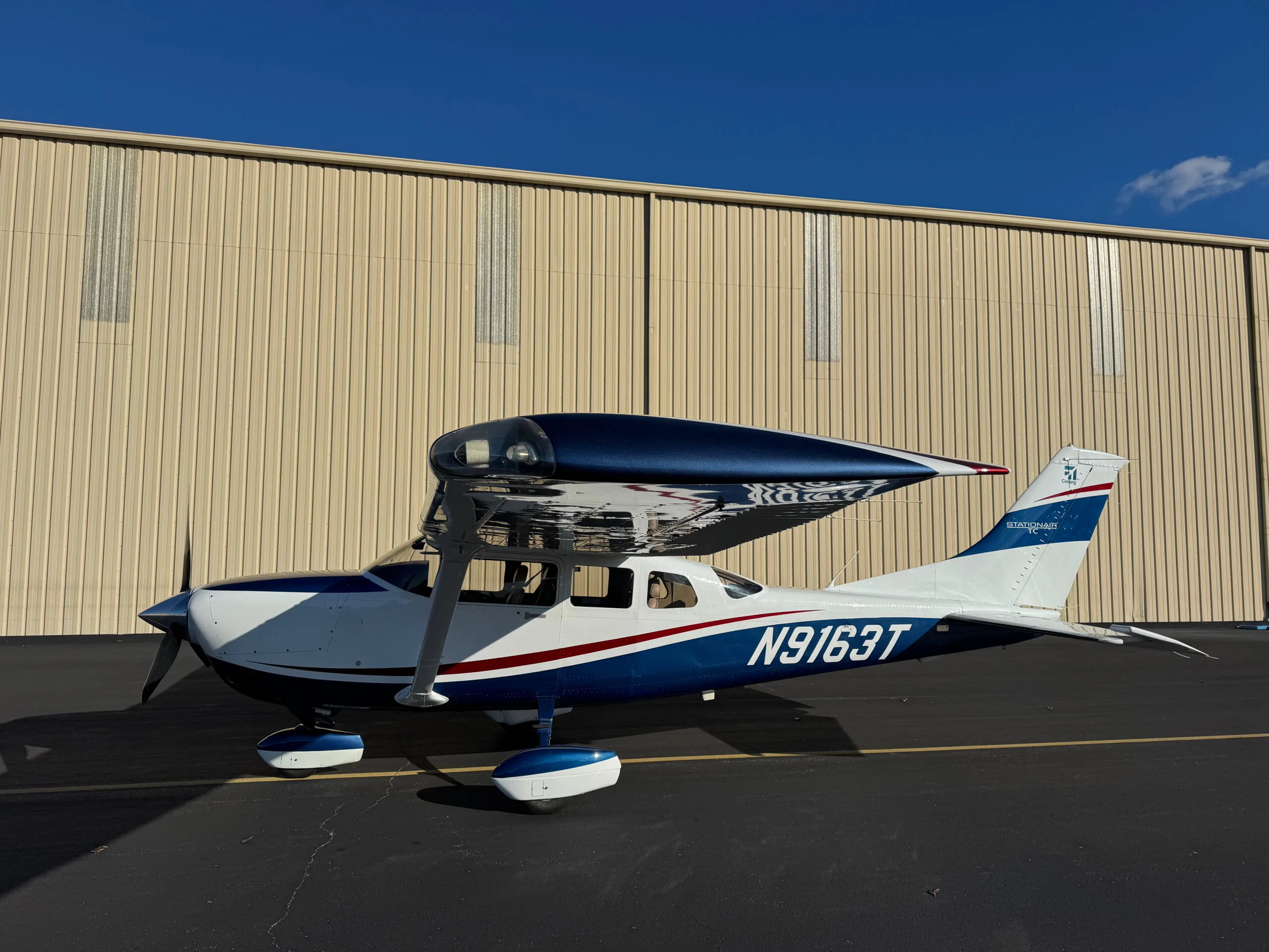 A white Cessna Stationair TC with blue and red accents is parked on an asphalt tarmac in front of a light-colored hangar under a clear blue sky.