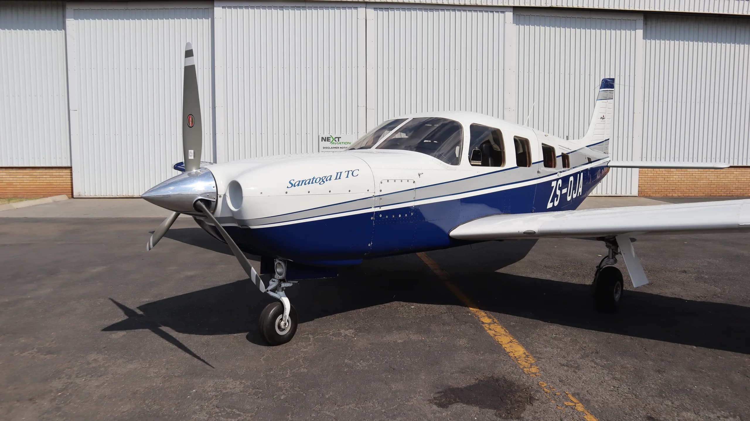 A white and blue Piper Saratoga II TC aircraft is parked on an asphalt in front of a white hangar.