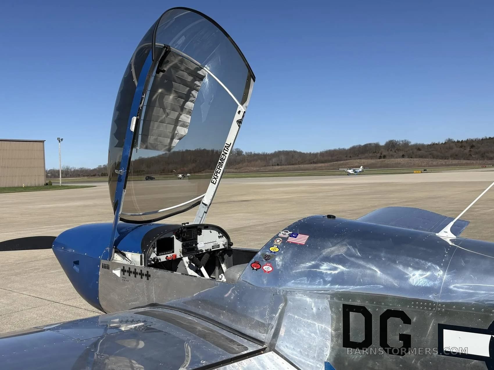 A blue and polished aluminum single-engine aircraft with its bubble canopy open, parked on an airfield.