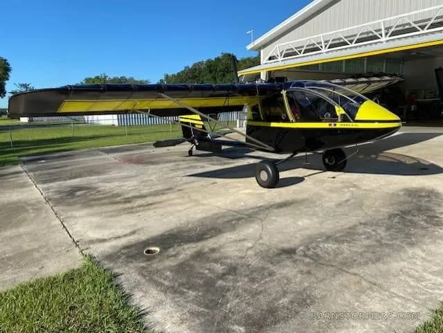 A black and yellow ultralight aircraft with a high wing, parked on a concrete pad in front of a hangar.