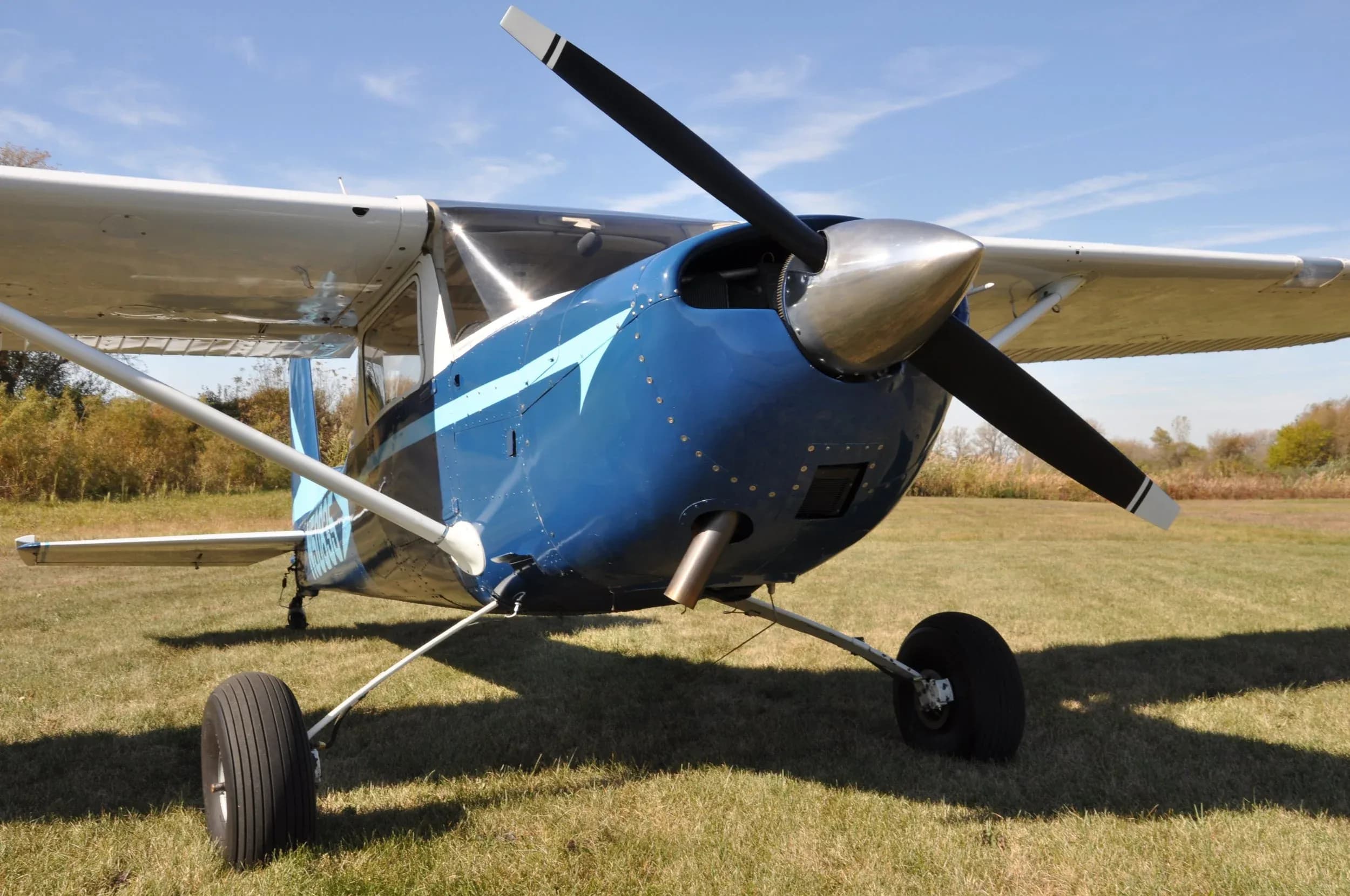 A close-up front view of a blue and white Cessna-like single-engine aircraft with a silver propeller spinner, parked on a grassy field.
