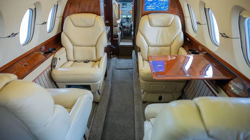 The private jet cabin interior looking forward, showing cream leather seats, a fold-out table, and a glimpse of the cockpit.