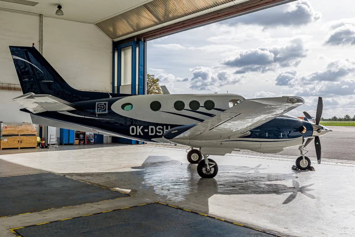 A white and blue Beechcraft King Air C90 twin-turboprop aircraft parked inside a hangar, with a cloudy sky visible outside.