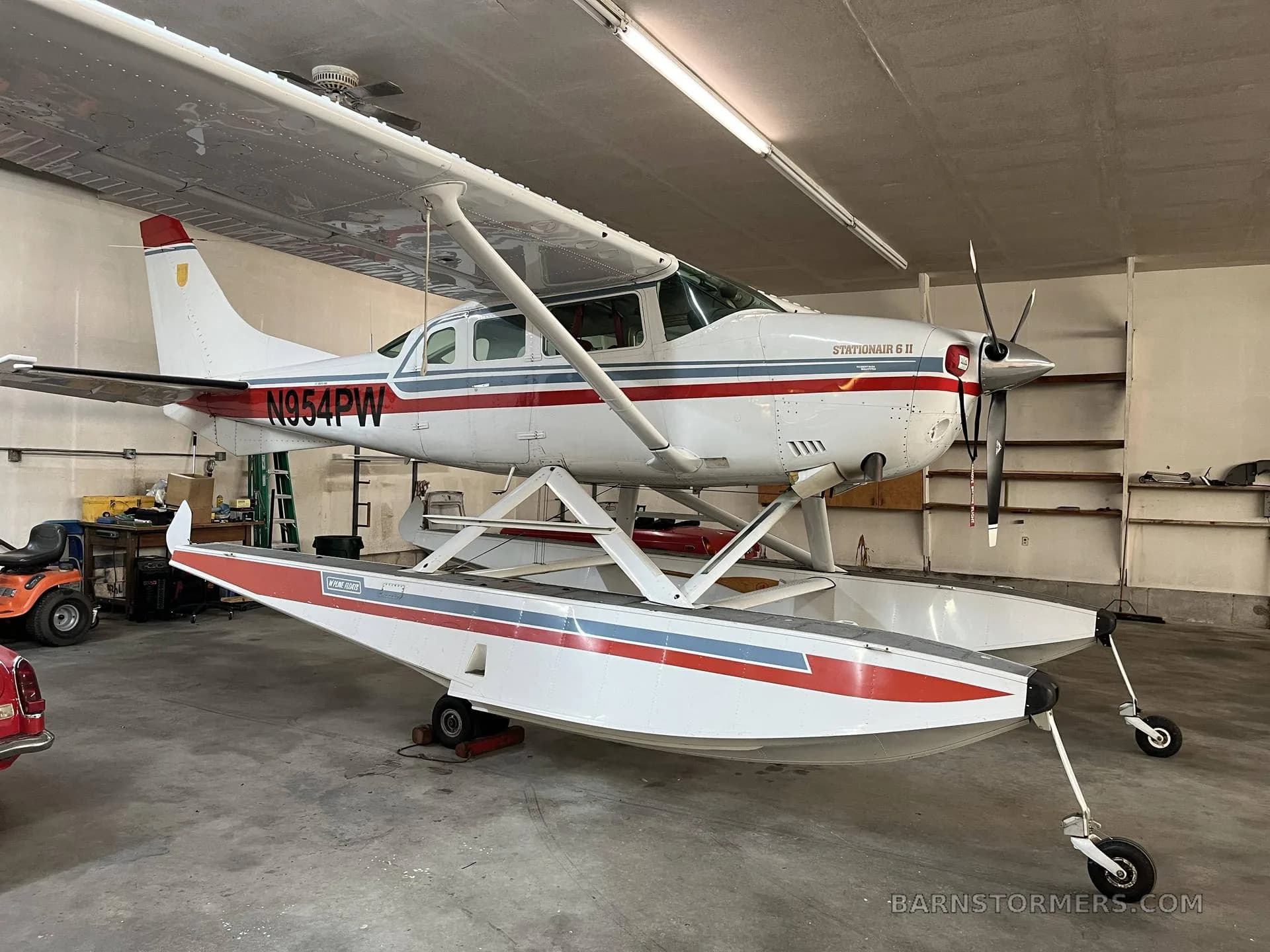 A white Cessna Stationair 6 II floatplane with red and blue stripes, registration N954PW, parked inside a hangar.
