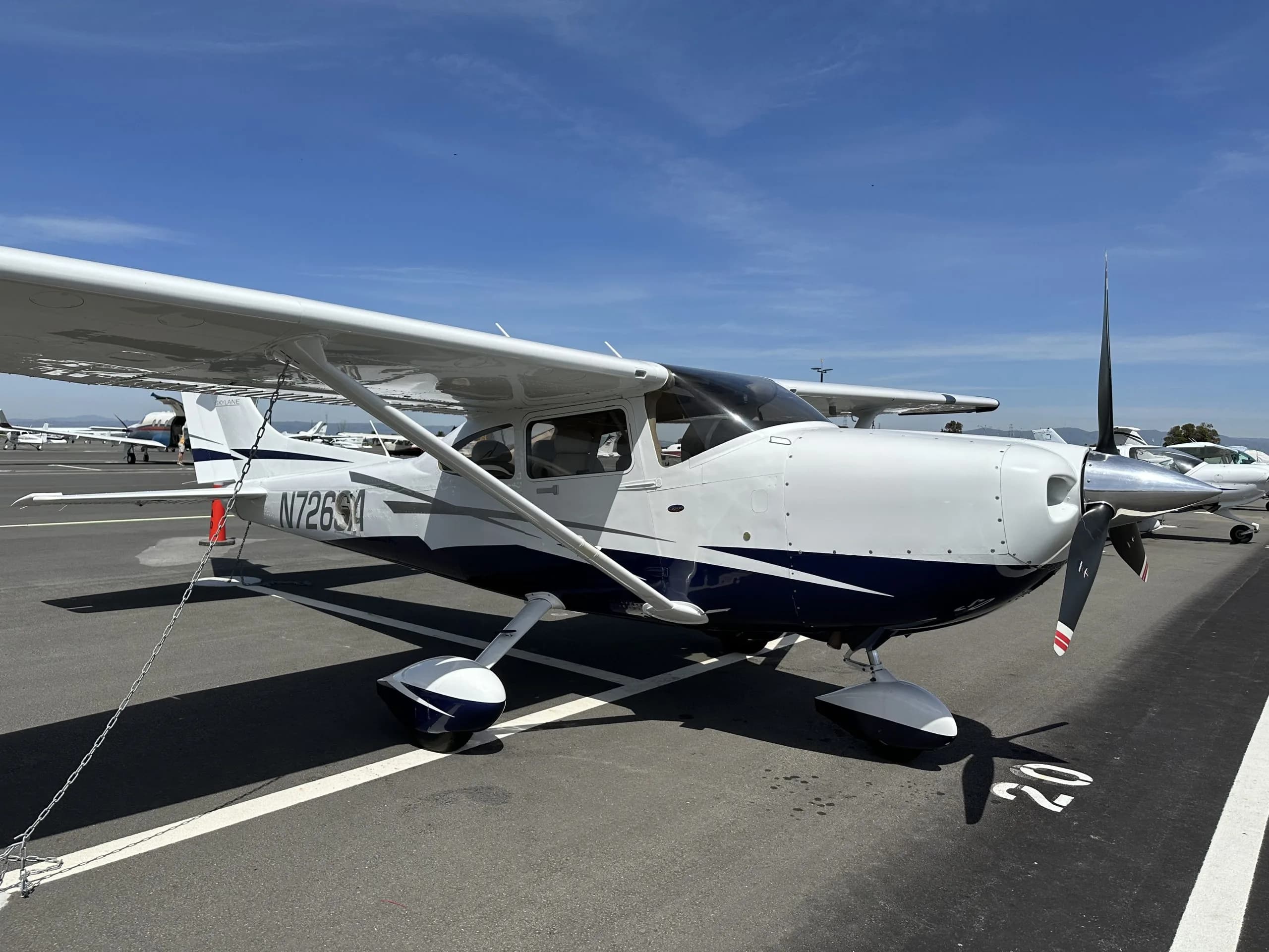 A white and blue Cessna 182 Skylane aircraft, N726SA, is parked on an airfield ramp under a clear blue sky, viewed from the front-right.