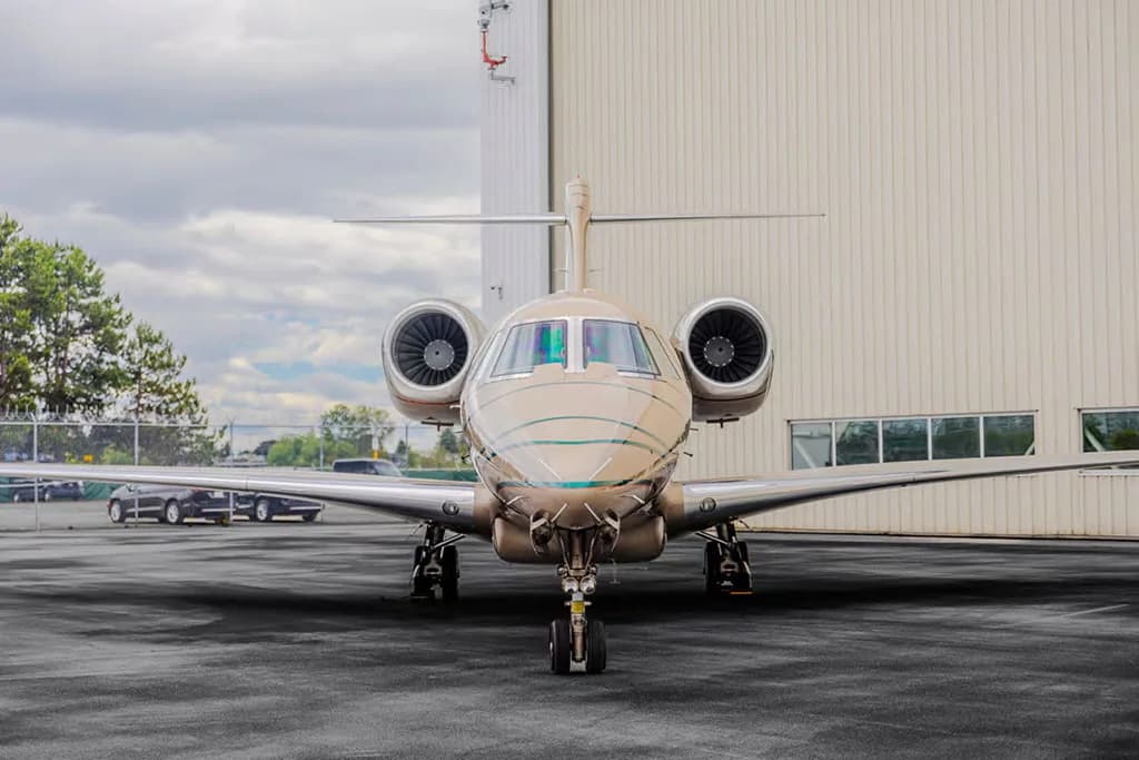 A head-on view of a beige business jet with green and brown stripes parked on an asphalt apron in front of a white hangar.